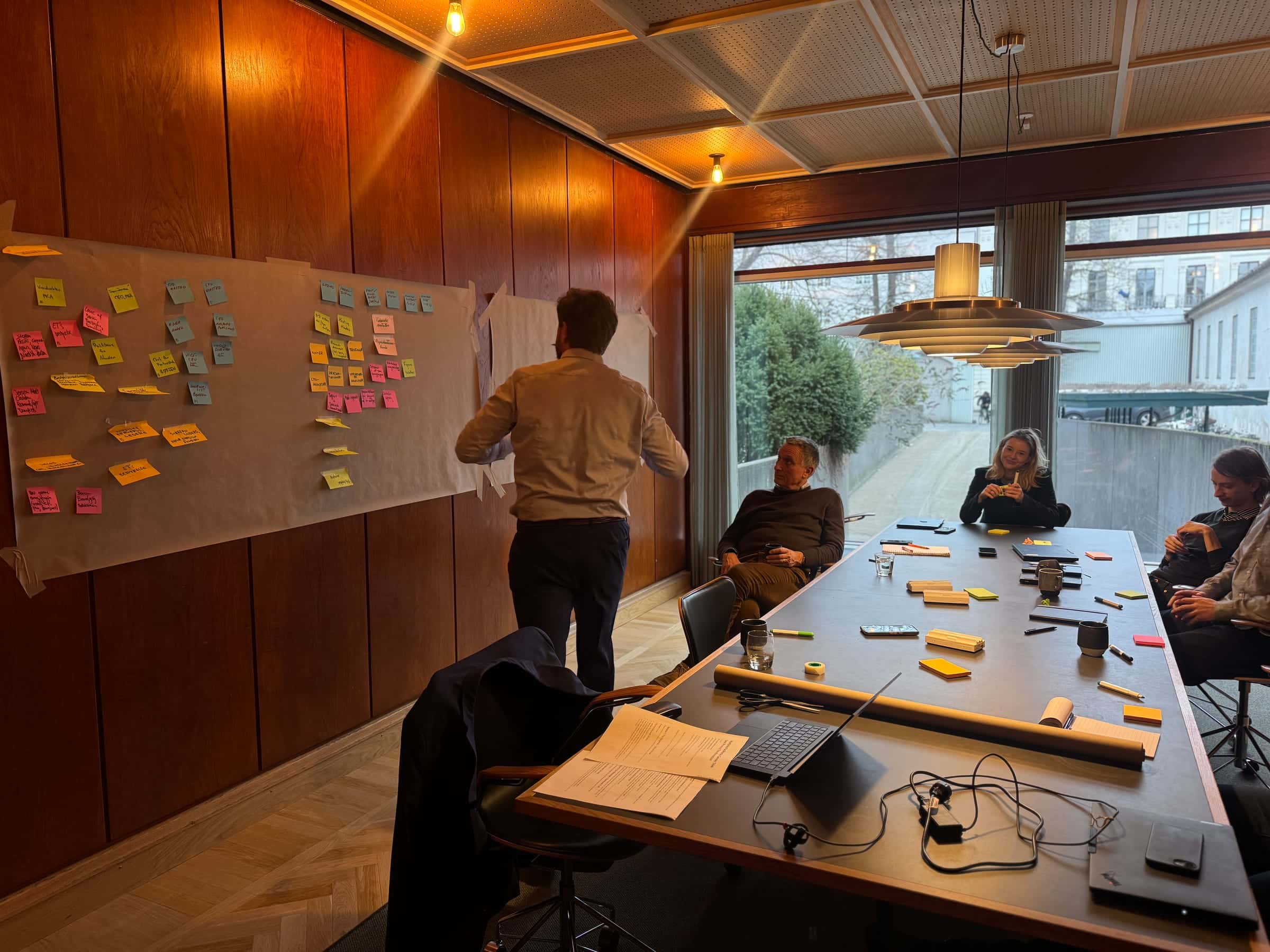 Simon facilitating a customer workshop, sticky-note canvas pinned to a wood-panelled wall, team seated around a long boardroom table.